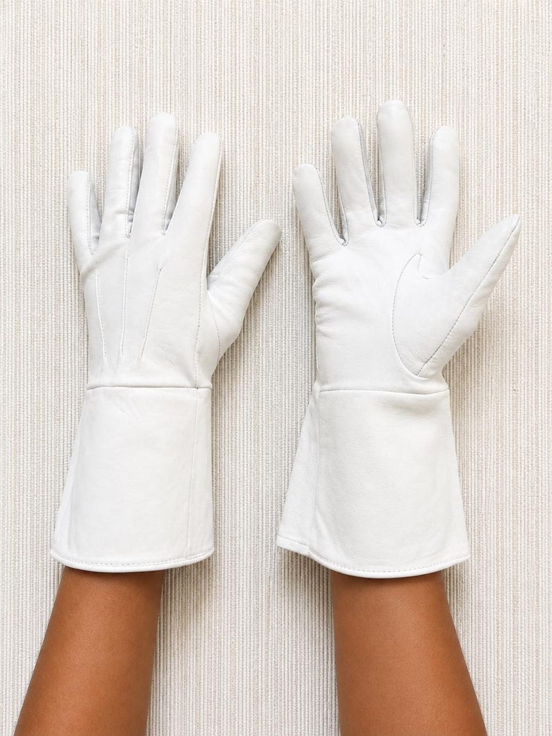 May include: A pair of white leather gloves featuring long, flared cuffs that extend past the wrist. The gloves have visible stitching details on the back of the hand and a smooth, matte finish, covering the fingers and palms completely.