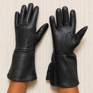 May include: A pair of black leather gloves featuring a smooth finish, five fingers, and an extended cuff design. The gloves have visible stitching details on the back of the hand and a structured thumb section, providing full coverage for the hands.