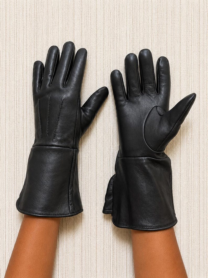 May include: A pair of black leather gloves featuring a smooth finish, five fingers, and an extended cuff design. The gloves have visible stitching details on the back of the hand and a structured thumb section, providing full coverage for the hands.