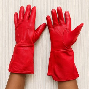 May include: A pair of vibrant red leather gloves featuring a smooth finish, five fingers, and an extended cuff design that reaches past the wrist. The gloves are constructed with visible stitching along the fingers and a structured, flared opening.