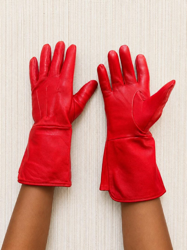 May include: A pair of vibrant red leather gloves featuring a smooth finish, five fingers, and an extended cuff design that reaches past the wrist. The gloves are constructed with visible stitching along the fingers and a structured, flared opening.