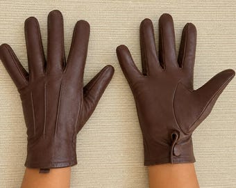 May include: A pair of dark brown leather gloves. The left glove shows the back with three vertical stitched lines. The right glove shows the palm side with a reinforced thumb panel and a small leather button closure at the wrist cuff.