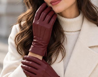 May include: A pair of deep burgundy leather gloves featuring three vertical stitched lines on the back of the hand, a smooth finish, and a straight cuff design. The gloves are worn over a cream-coloured ribbed turtleneck sweater and a coat.