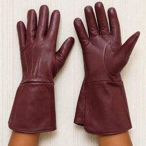 May include: A pair of deep burgundy leather gloves featuring a smooth finish, five fingers, and an extended cuff design that reaches past the wrist. The gloves have visible stitching details along the back of the hand and a structured thumb section.