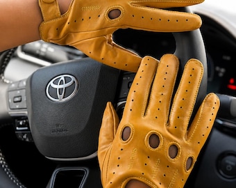 May include: A pair of mustard yellow leather driving gloves featuring perforated detailing, knuckle cutouts, and a snap button closure at the wrist. The gloves are designed with a classic style, showcasing visible stitching and a smooth texture.