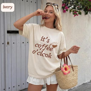 May include: Ivory-colored t-shirt with the text "it's coffee o'clcok" in brown. The shirt features a coffee pot graphic. A straw tote bag with a pink flower is held in the hand. The person is holding cherries.