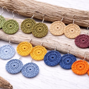 Può includere: Una collezione di orecchini all'uncinetto fatti a mano in vari colori, tra cui verde, blu, giallo, arancione, beige e bordeaux. Ogni orecchino presenta un design circolare con un foro centrale e ganci argentati. Presentati su legno.