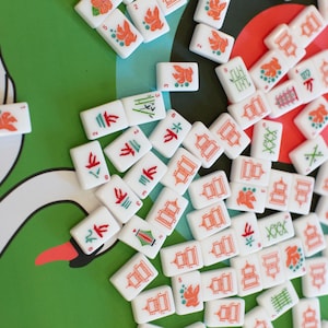 Puede incluir: Un conjunto de fichas de Mahjong blancas con símbolos y caracteres rojos, verdes y naranjas esparcidos sobre una superficie verde con una ilustración de cisne. Las fichas presentan varios diseños, incluyendo flores, pagodas y bambú.