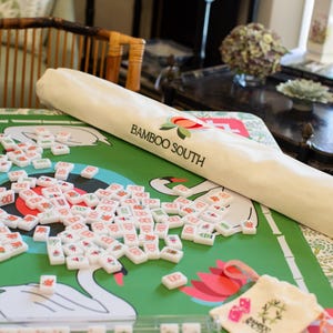 May include: A green mahjong game mat with swan and lotus flower designs. The mat is partially covered by a rolled-up beige carrying case with the text "Bamboo South". Scattered white mahjong tiles with red, green, and orange symbols are on the mat.