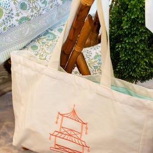 May include: Cream-colored tote bag with a coral-colored pagoda design. The bag has cream-colored straps and a light blue interior. The bag is resting on a wooden chair with a green plant in the background. The background also includes a floral patterned fabric.