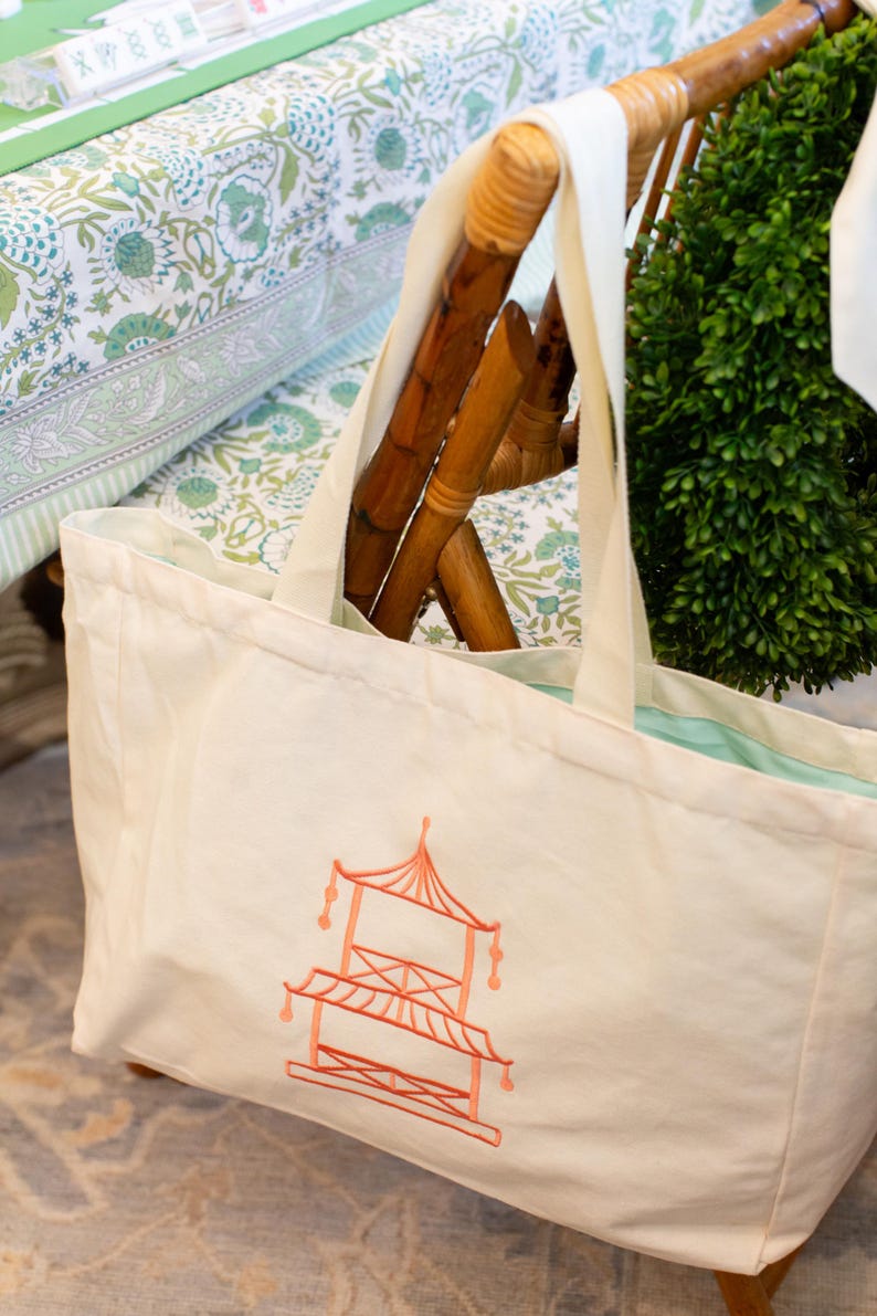 May include: Cream-colored tote bag with a coral-colored pagoda design. The bag has cream-colored straps and a light blue interior. The bag is resting on a wooden chair with a green plant in the background. The background also includes a floral patterned fabric.