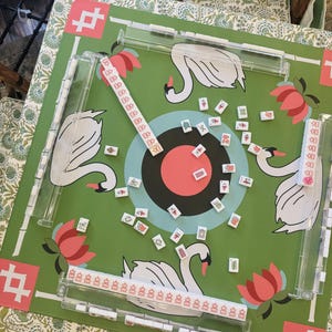 May include: A green Mahjong game set with a swan and lotus flower design. The game board features a target in the center and is surrounded by clear plastic tile holders. The tiles are scattered across the board.