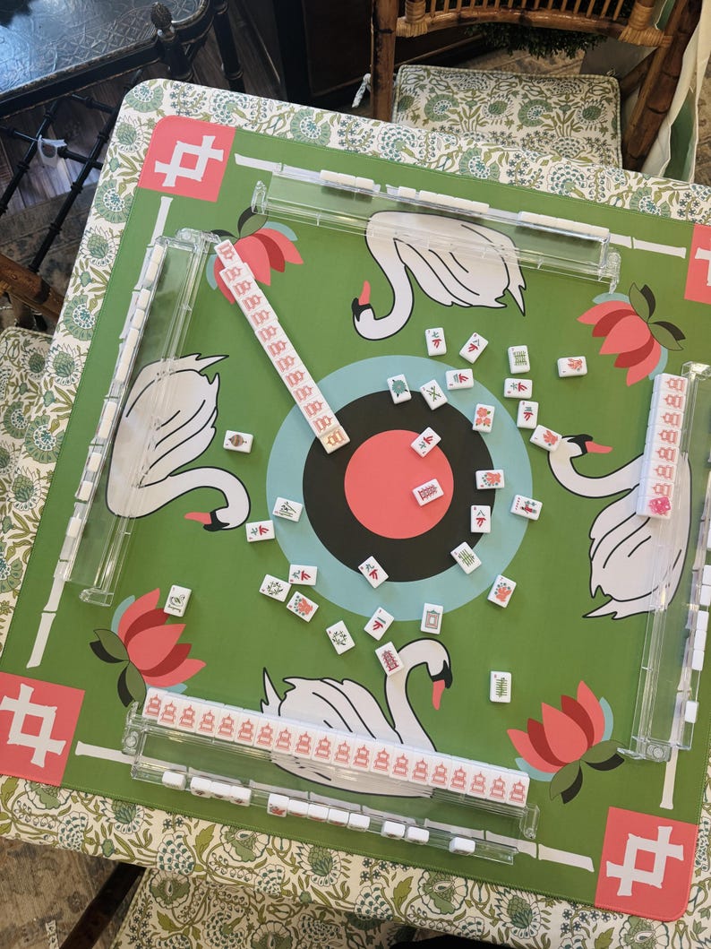 May include: A green Mahjong game set with a swan and lotus flower design. The game board features a target in the center and is surrounded by clear plastic tile holders. The tiles are scattered across the board.
