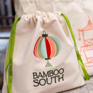 May include: Off-white drawstring bag with a colorful embroidered lantern design. The lantern features red, orange, green, and blue sections. The text "BAMBOO SOUTH" is embroidered below the lantern in dark green. Green ribbon ties.