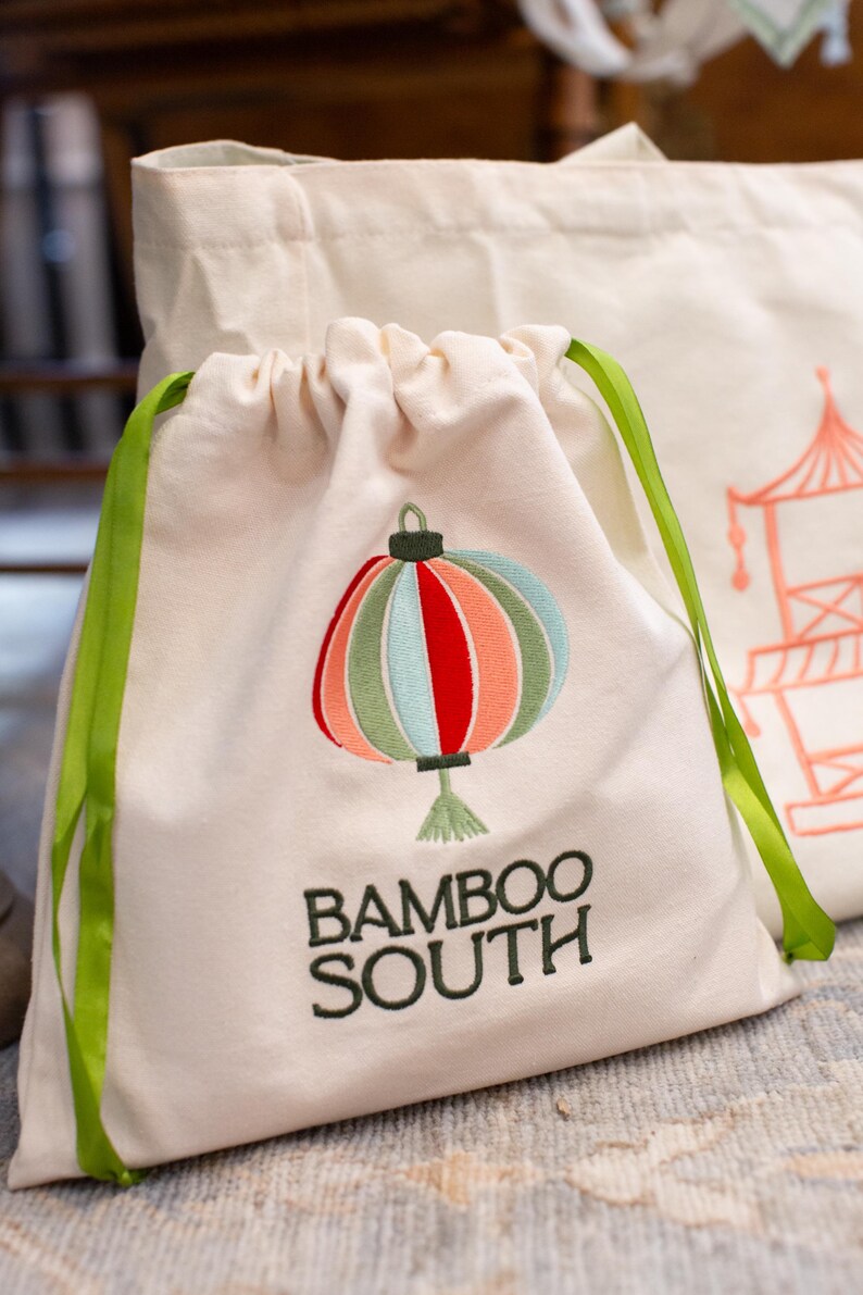 May include: Off-white drawstring bag with a colorful embroidered lantern design. The lantern features red, orange, green, and blue sections. The text "BAMBOO SOUTH" is embroidered below the lantern in dark green. Green ribbon ties.