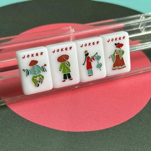 May include: Four white Mahjong tiles with the word "JOKER" printed on each. Each tile features a different illustration of a person in a costume. The tiles are in a clear plastic holder, set against a background of a pink and green circular design.