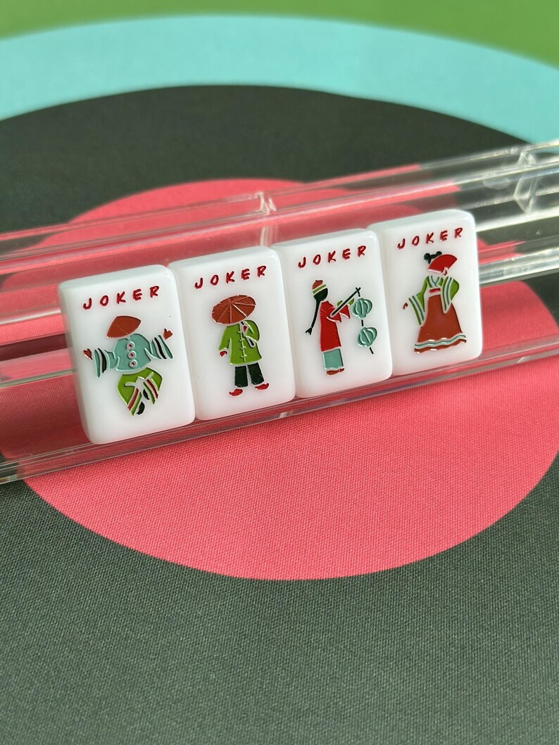 May include: Four white Mahjong tiles with the word "JOKER" printed on each. Each tile features a different illustration of a person in a costume. The tiles are in a clear plastic holder, set against a background of a pink and green circular design.