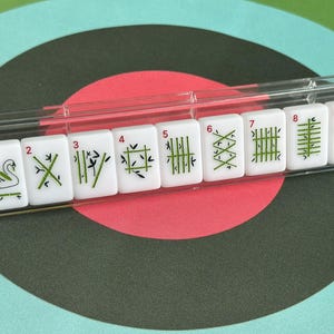 May include: A set of white Mahjong tiles with green and black symbols, numbered 1 through 9. The tiles are displayed in a clear plastic holder against a background of concentric circles in teal, red, and black.