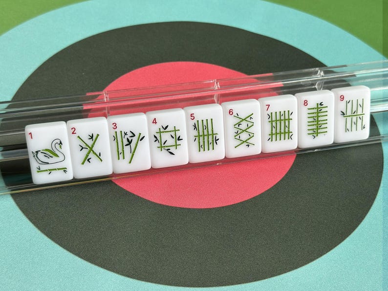 May include: A set of white Mahjong tiles with green and black symbols, numbered 1 through 9. The tiles are displayed in a clear plastic holder against a background of concentric circles in teal, red, and black.