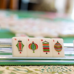 May include: Close-up of Mahjong tiles with colorful lantern designs and the word "JOKER". The tiles are arranged on a green surface, suggesting a game in progress. The image is well-lit and focuses on the details of the tiles.