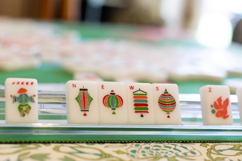 May include: Close-up of Mahjong tiles with colorful lantern designs and the word "JOKER". The tiles are arranged on a green surface, suggesting a game in progress. The image is well-lit and focuses on the details of the tiles.