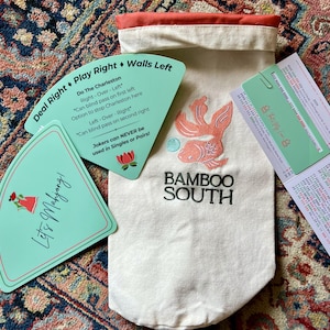 May include: A Mahjong game set featuring a canvas bag with an embroidered orange fish and the text "BAMBOO SOUTH." Included are teal game cards with instructions and the text "Let's Mahjong!" and "Deal Right Play Right Walls Left."