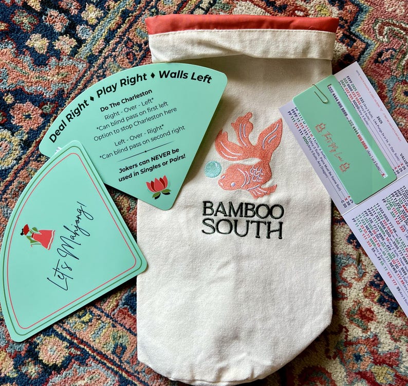May include: A Mahjong game set featuring a canvas bag with an embroidered orange fish and the text "BAMBOO SOUTH." Included are teal game cards with instructions and the text "Let's Mahjong!" and "Deal Right Play Right Walls Left."