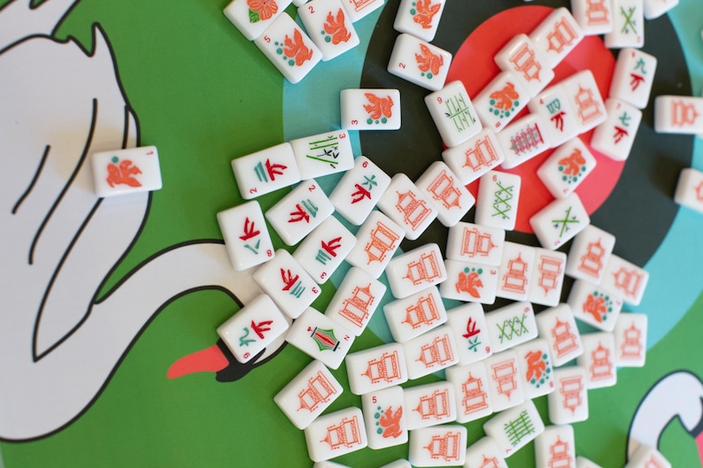 May include: A collection of white Mahjong tiles with red, green, and orange symbols and characters scattered on a green surface with a swan illustration. The tiles feature various designs, including flowers, pagodas, and bamboo.
