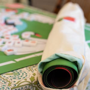 May include: A rolled-up green and black game mat with red accents is partially visible inside a cream-colored drawstring bag. The game mat is on a floral patterned surface, with a partially visible game board and game pieces in the background.
