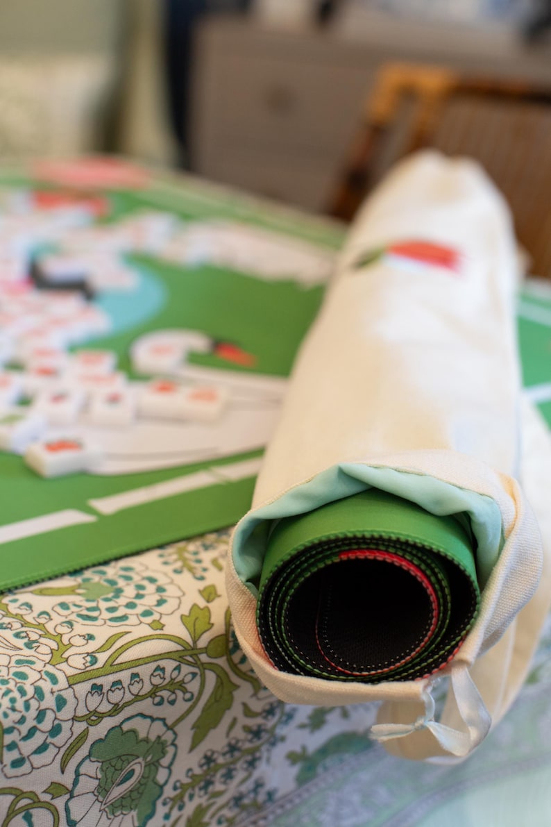 May include: A rolled-up green and black game mat with red accents is partially visible inside a cream-colored drawstring bag. The game mat is on a floral patterned surface, with a partially visible game board and game pieces in the background.