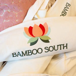 May include: Off-white fabric with an embroidered lotus flower in shades of red, peach, green, and blue. The text "BAMBOO SOUTH" is embroidered below the flower in dark green. Other off-white fabric items are in the background.