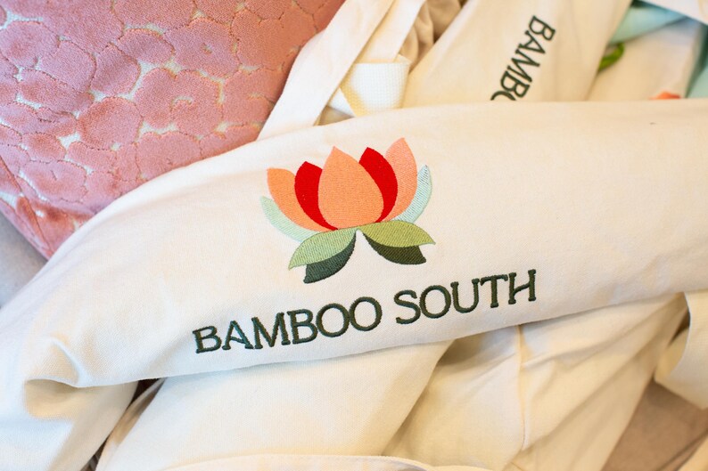 May include: Off-white fabric with an embroidered lotus flower in shades of red, peach, green, and blue. The text "BAMBOO SOUTH" is embroidered below the flower in dark green. Other off-white fabric items are in the background.