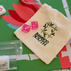 May include: A close-up of a Mahjong set with pink dice and a cream-colored bag. The bag has the text "Bamboo South" and a green bamboo design. The background is a green and red floral pattern.