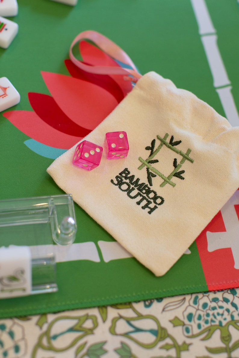 May include: A close-up of a Mahjong set with pink dice and a cream-colored bag. The bag has the text "Bamboo South" and a green bamboo design. The background is a green and red floral pattern.