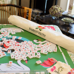 May include: A green mahjong game mat with a swan design and the text "Bamboo South". The image shows a set of mahjong tiles scattered on the mat, along with a matching carrying case and a rolled-up mat.