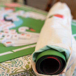May include: A rolled-up green and black game mat with red accents is partially visible inside a cream-colored drawstring bag. The game mat is on a floral patterned surface, with a partially visible game board and game pieces in the background.