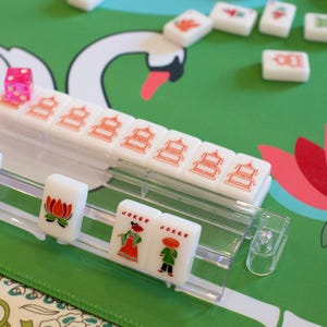 May include: A close-up of a Mahjong set with white tiles featuring red and orange symbols. The set includes a pink die and tiles with "Joker" text. The game is set on a green surface with a swan design.
