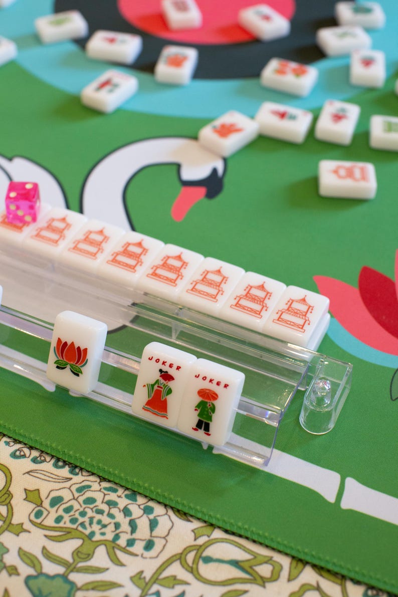 May include: A close-up of a Mahjong set with white tiles featuring red and orange symbols. The set includes a pink die and tiles with "Joker" text. The game is set on a green surface with a swan design.