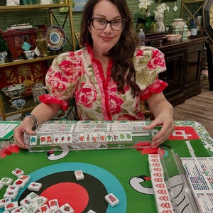 May include: A woman wearing glasses and a floral top is seated at a green table playing Mahjong. The table is covered with a green cloth with a red circle design. Mahjong tiles are scattered on the table, and a clear plastic tile holder is in front of the woman.
