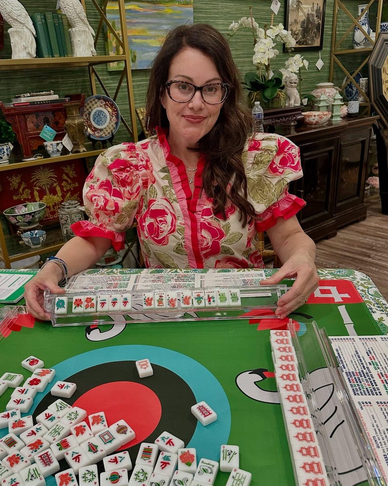 May include: A woman wearing glasses and a floral top is seated at a green table playing Mahjong. The table is covered with a green cloth with a red circle design. Mahjong tiles are scattered on the table, and a clear plastic tile holder is in front of the woman.