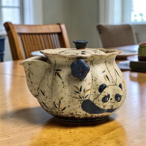 Puede incluir: Tetera de cerámica con diseño de panda. La tetera es beige con detalles en negro y azul, con una ilustración de oso panda y tallos de bambú. Tiene un pico, un asa y una tapa con un botón azul oscuro.