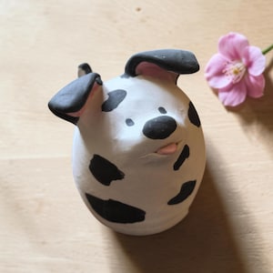 Op de afbeelding: Handgemaakte keramische hondenfiguur met zwarte vlekken en oren, een zwarte neus en een roze tong. De hond is wit met zwarte vlekken en heeft een speelse uitdrukking. Op de achtergrond staat een roze bloem.