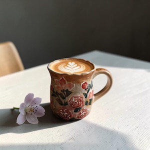 May include: A ceramic mug with a floral pattern, filled with a latte. The mug is beige with red and pink roses. A single pink flower lies on the table beside the mug. The latte art has a leaf design.