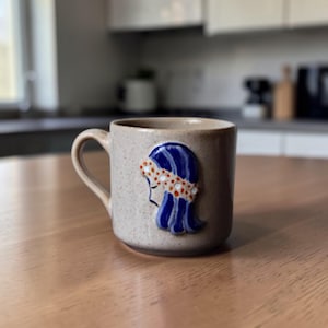 Può includere: Una tazza in ceramica beige macchiettata con manico, con un design decorativo blu e bianco di un profilo femminile. La donna ha i capelli blu e una fascia bianca con pois rossi. La tazza è su un tavolo di legno.