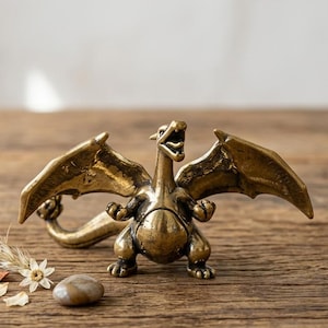 Puede incluir: Una figura de dragón de bronce con las alas extendidas y la boca abierta. El dragón está posado sobre una superficie de madera, acompañado de flores secas y una pequeña piedra. La figura tiene detalles intrincados y un acabado metálico.