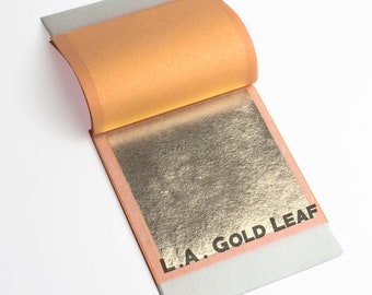 May include: A partially opened book of gold leaf sheets. The gold leaf is a bright, metallic gold color. The book has a light orange cover and the text "L.A. GOLD LEAF" is printed on the bottom right corner.