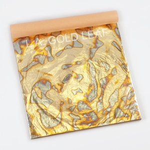 May include: A sheet of gold leaf with a swirling, marbled pattern in shades of gold and silver. The leaf is rolled up and labeled "LA GOLD LEAF".