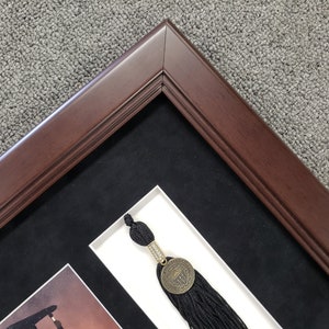 May include: A dark brown wooden picture frame with a black mat and a white inner mat. A black tassel with a gold medallion is visible in the frame.