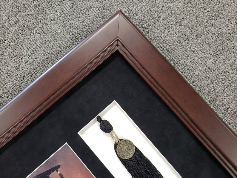 May include: A dark brown wooden picture frame with a black mat and a white inner mat. A black tassel with a gold medallion is visible in the frame.