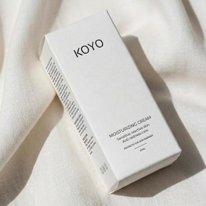 Op de afbeelding: Een witte rechthoekige doos met KOYO vochtinbrengende crème. De doos heeft de merknaam KOYO in zwarte letters. De tekst "MOISTURIZING CREAM" en andere details staan op de doos. Het product is ontworpen voor de gevoelige, reactieve huid en anti-roodheid.